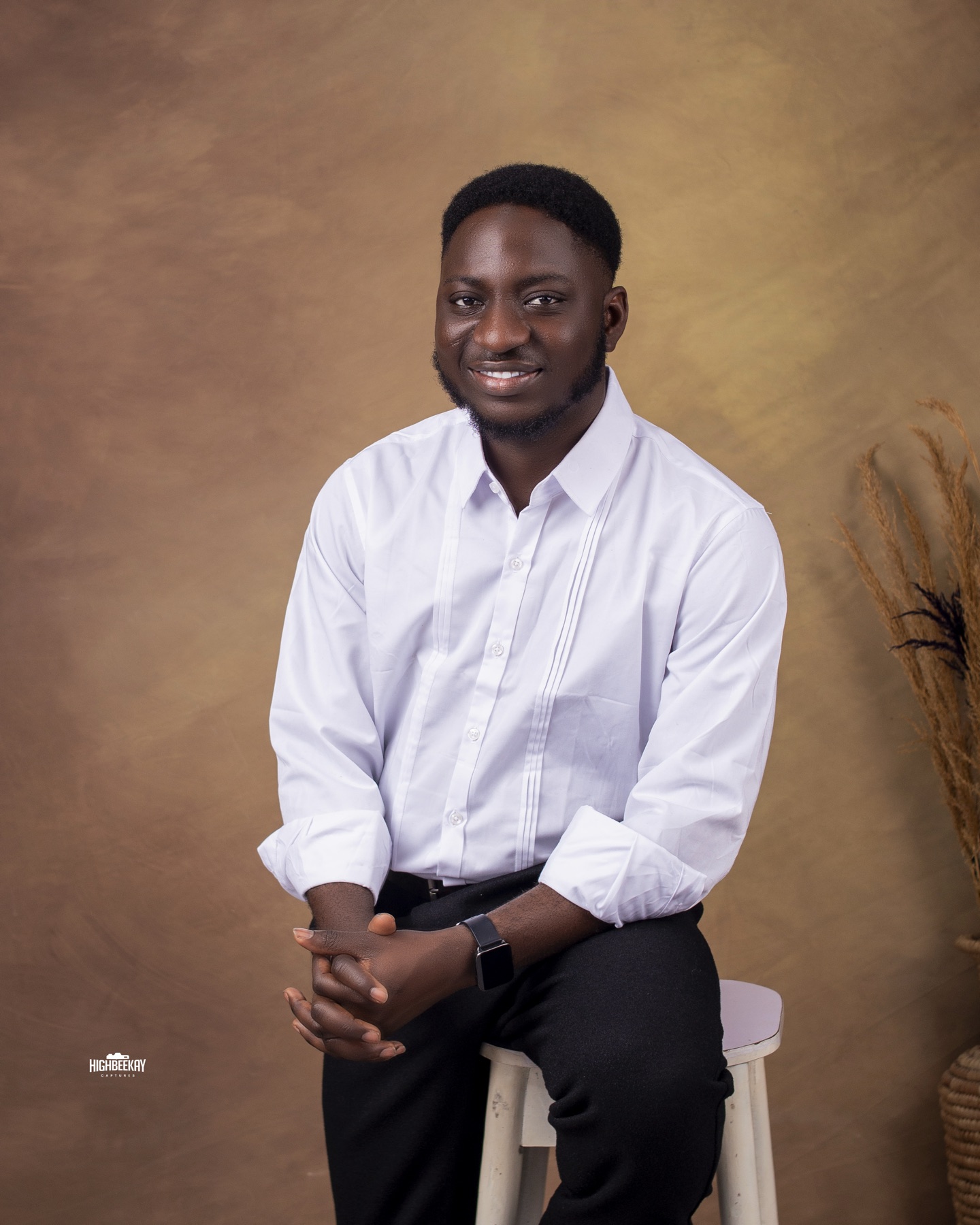 Michael Akintayo seated in a white shirt against a warm brown studio backdrop